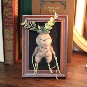 May include: A brown felt creature with green leaves growing from its head is framed in a brown wooden frame. The creature has a sad expression and long, thin, green roots.
