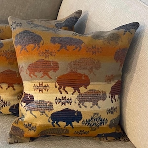 May include: Decorative throw pillows with a bison pattern. The pillows feature a repeating pattern of bison in shades of brown, orange, and blue against a tan and orange striped background. The pillows are on a light-colored sofa.