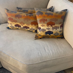 May include: Three throw pillows with a beige background and a pattern of brown and blue buffalo silhouettes. The pillows are on a light beige couch.