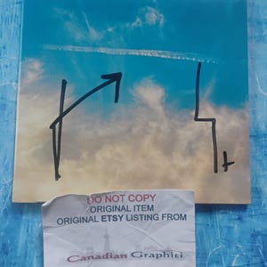 May include: A square print featuring a sky with clouds and black line art. The print has an arrow and a lightning bolt design. A sticker at the bottom reads "DO NOT COPY ORIGINAL ITEM ORIGINAL ETSY LISTING FROM Canadian Graphiti."