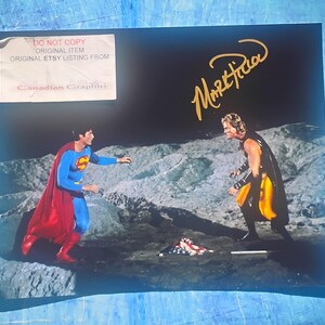 Puede incluir: Fotografía de Superman con un traje rojo y azul con capa roja, frente a un personaje con un atuendo negro y amarillo. El fondo es un paisaje oscuro y rocoso. La foto tiene una firma dorada. Una pegatina dice "DO NOT COPY ORIGINAL ITEM ORIGINAL ETSY LISTING FROM Canadian Graphiti."
