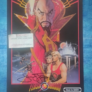 以下が含まれることがあります： 映画「フラッシュ・ゴードン」のヴィンテージ映画ポスター。長いひげを生やした厳しい顔の男性が中心に描かれ、他のキャラクターに囲まれています。ポスターには「Pathetic Earthlings... Who Can Save You Now?」と「Music by QUEEN」の文字が含まれています。