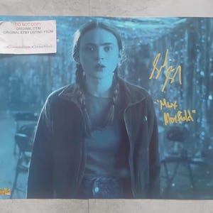 May include: A poster featuring a person with braided hair, wearing a dark jacket and jeans. The poster has a blue and white background. The person's signature and the name "Max Mayfield" are in gold ink. A sticker is in the upper left corner.