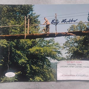 May include: A signed photograph featuring a man standing on a wooden bridge, surrounded by lush green trees. The photograph has the signature "Billy Halter" in blue ink. A sticker with text is visible in the lower right corner.