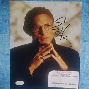 Könnte beinhalten: Ein glänzendes Foto mit einem Porträt von Ed Harris, der eine dunkle Mütze und eine Brille trägt, die Hände gefaltet mit Ringen an den Fingern. Das Bild enthält eine Unterschrift in schwarzer Tinte, einen Echtheitsaufkleber und ein weißes Etikett.