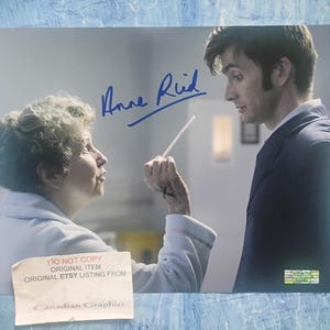 May include: A glossy photograph featuring an older person in a white robe and a man in a dark suit, with the blue ink signature of Anne Reid. A small white paper label at the bottom reads DO NOT COPY ORIGINAL ITEM ORIGINAL ETSY LISTING FROM.