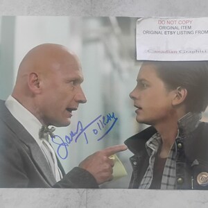 May include: A signed photograph featuring two people in a scene. One person, with a bald head, is wearing a suit and pointing. The other person is wearing a jacket over a plaid shirt. The photo includes the text "DO NOT COPY ORIGINAL ITEM ORIGINAL ETSY LISTING FROM Canadian Graphiti".