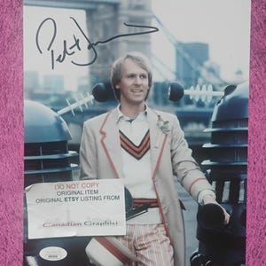 May include: A glossy photograph featuring a person in a beige jacket with red trim, a white sweater with red and black stripes, and striped trousers, standing between two dark blue robotic figures. A black ink signature is visible at the top.