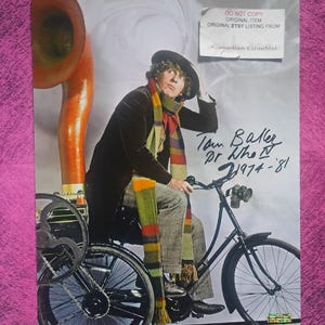 May include: A photograph of a person in a dark coat, grey trousers, and a long multi-colored knit scarf, sitting on a black bicycle with a large orange horn attached. The image features a signature and the text Tom Baker Dr Who IV 1974-81.