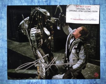 Może przedstawiać: Podpisany wydruk przedstawiający scenę z filmu Predator. Obraz przedstawia Predatora w zbroi naprzeciwko mężczyzny w szarej kurtce. Wydruk ma białą naklejkę z napisem "DO NOT COPY ORIGINAL ITEM ORIGINAL ETSY LISTING FROM Canadian Graphiti."
