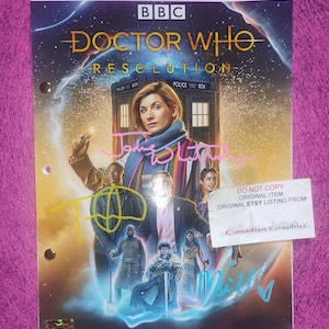 Pode incluir: Uma capa de roteiro de Doctor Who Resolution com o logotipo da BBC, o título e uma ilustração do elenco com a TARDIS. A capa inclui três autógrafos distintos em tinta rosa, amarela e azul, junto com uma etiqueta DO NOT COPY.