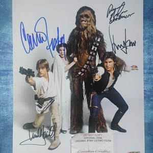 May include: A Star Wars cast photo featuring Luke Skywalker, Princess Leia, Chewbacca, and Han Solo, each holding a blaster. The photo includes autographs in blue ink. A white label at the bottom reads "DO NOT COPY ORIGINAL ITEM ORIGINAL ETSY LISTING FROM Canadian Graphiti."