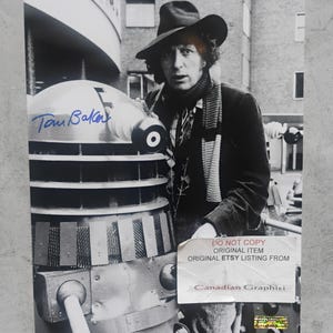 May include: A black and white photograph featuring Tom Baker wearing a wide-brimmed hat and a long scarf, standing next to a Dalek. The image includes a blue ink signature and a white label with the text DO NOT COPY ORIGINAL ITEM CANADIAN GRAPHITI.