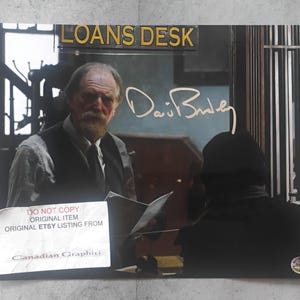 Peut inclure: Une photographie rectangulaire montrant un homme barbu portant un gilet sombre derrière un comptoir marqué LOANS DESK. L'image comprend une signature dorée, une étiquette blanche avec le texte DO NOT COPY et un petit autocollant circulaire.