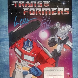Könnte beinhalten: Ein Poster mit dem The Transformers-Logo oben, Optimus Prime im Vordergrund und Megatron im Hintergrund vor einem sternenklaren violetten Weltraumhintergrund. Das Poster enthält zwei Unterschriften in blauer Tinte und einen weißen Aufkleber unten.