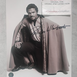 May include: Black and white photograph of Billy Dee Williams, signed. He is wearing a light brown cape and a dark shirt. The photo includes text that reads "DO NOT COPY ORIGINAL ITEM ORIGINAL ETSY LISTING FROM Canadian Graphiti."