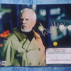 May include: A signed photograph featuring a man with white hair and a beard, wearing a tan coat with a green collar. The background is blurred with city lights. The photo includes text that reads "DO NOT COPY ORIGINAL ITEM ORIGINAL ETSY LISTING FROM Canadian Graphiti."