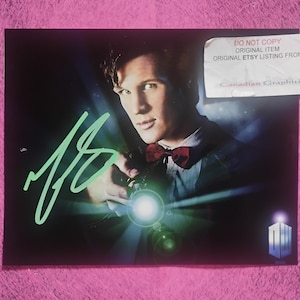 May include: A rectangular photograph featuring a man in a dark suit and red bow tie holding a glowing device, with a light green autograph across the left side. A white sticker in the top right corner reads DO NOT COPY ORIGINAL ITEM.