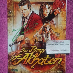 Pode incluir: Uma capa de roteiro impressa para The Rings of Akhaten com um homem de jaqueta vermelha, uma mulher segurando uma folha vermelha e três figuras mascaradas. O título está em letras amarelas, com um autógrafo verde e créditos de produção.