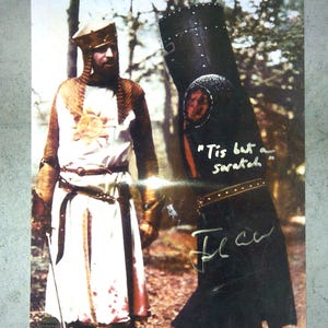 May include: A photographic print featuring a knight in white and gold armor standing next to a black-armored knight. The black knight has the text "Tis but a scratch" and a signature written on the surface. The white knight wears a sun emblem.