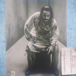 May include: Black and white print of a person in a white outfit with makeup, seated on a dark box. The person has long hair and a painted mustache. A sticker with text is in the lower right corner.