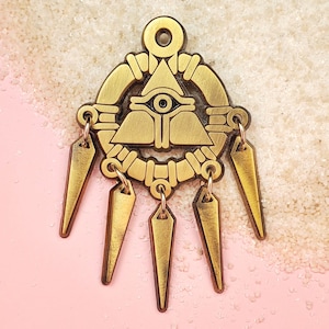 May include: Gold-toned pendant featuring an eye within a triangle, surrounded by a circular design with intricate details. The pendant hangs from five dangling chains with pointed ends.