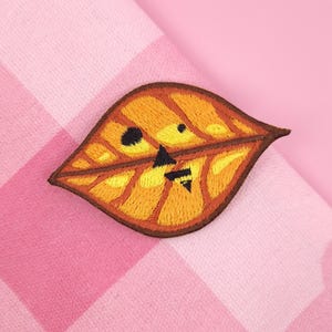 May include: Embroidered leaf patch in shades of orange, yellow, and brown, with a dark brown border. The leaf has a stylized face with black accents. The patch is set against a pink and white checkered background.