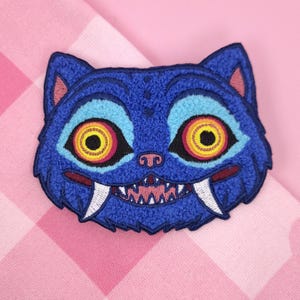 Könnte beinhalten: Ein blauer, gestickter Aufnäher in Form eines Katzenkopfes. Die Katze hat große, runde Augen mit gelben und roten Iris, eine rosa Nase und scharfe, weiße Fangzähne. Auf einem rosa-weiß karierten Hintergrund.