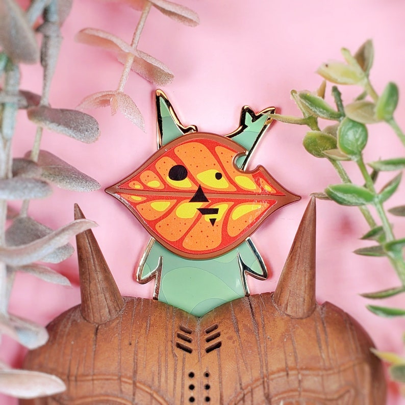 May include: A gold-tone enamel pin featuring a cartoon character with a green body and a large orange and yellow leaf-shaped head. The character is standing on a wooden base.
