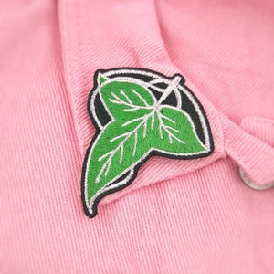 May include: A close-up of a pink fabric with a green leaf patch. The patch is embroidered with a detailed green leaf design, outlined in black and white. The fabric appears to be a textured cotton or similar material.