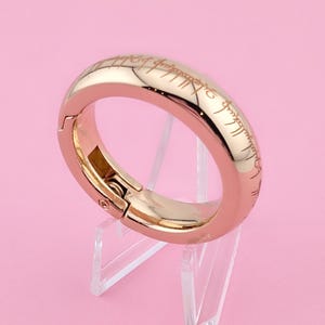 Puede incluir: Un anillo de oro rosa con una inscripción en escritura élfica. El anillo está abierto y tiene un cierre con bisagra.
