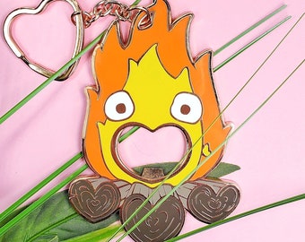 May include: A gold-colored metal bottle opener shaped like a cartoon fire spirit with a large, round, open mouth. The fire spirit is sitting on a bed of logs with heart-shaped details.