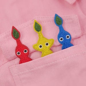 May include: Three colourful Pikmin-themed felt appliques, red, yellow, and blue, are tucked into the pocket of a pink shirt. Each applique features a leaf-shaped top and two white eyes. The shirt's pocket is visible, with the appliques arranged in a row.
