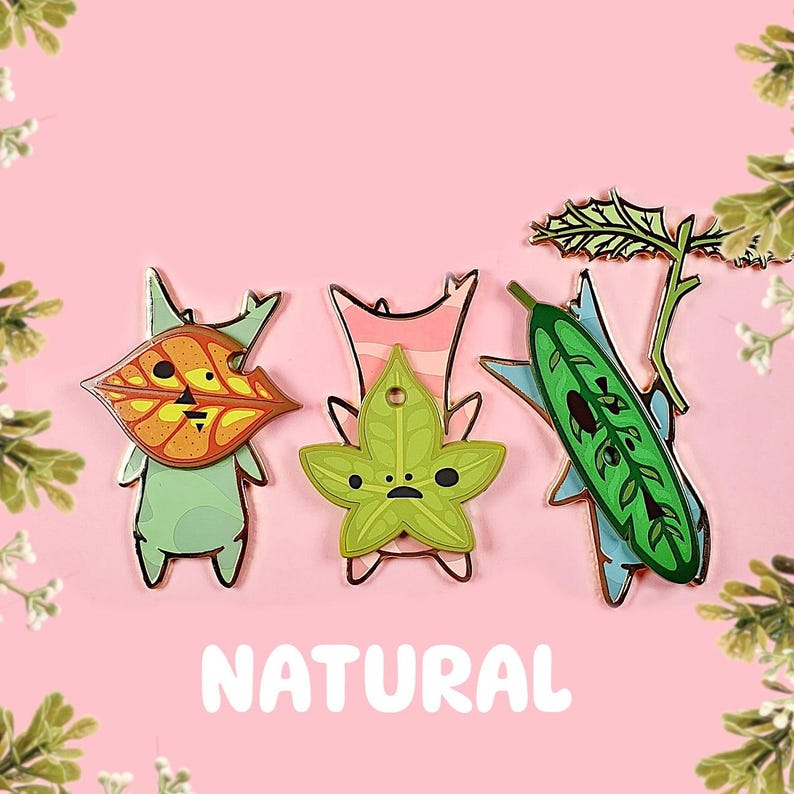 May include: Four enamel pins featuring cartoon characters with leaf-like features. The characters are green, pink, green, and blue. The pins are arranged in a row on a pink background. The word "NATURAL" is written in white letters below the pins.