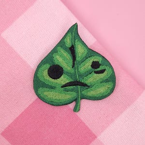 May include: Embroidered green leaf patch with a sad face design. The leaf has a detailed texture and a black, stitched face with two eyes and a downturned mouth. The patch is on a pink and white checkered background.