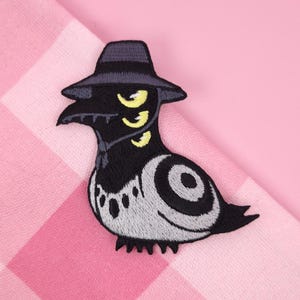 Peut inclure: Écusson brodé représentant un oiseau noir stylisé portant un chapeau fedora. L'oiseau a trois yeux jaunes et un corps gris et blanc avec un motif en spirale. L'écusson est sur un fond à carreaux roses et blancs.