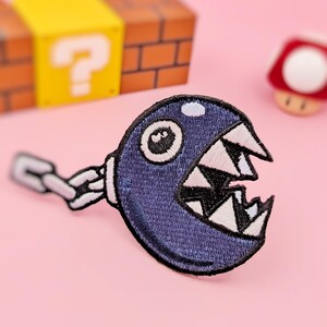 Puede incluir: Un parche bordado azul y blanco de un personaje de la serie de videojuegos Super Mario Bros. El personaje es un Chain Chomp con dientes afilados y un solo ojo. El parche está unido a una cadena.