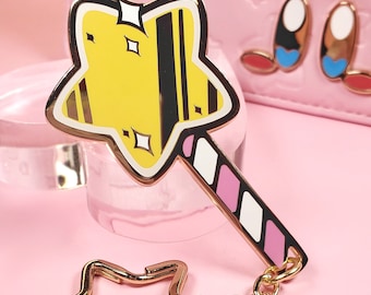 May include: Gold-tone keychain featuring a star-shaped wand with a pink and white striped handle. The star is yellow with black stripes and glitter accents.