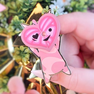 May include: Pink enamel pin with a heart-shaped head and a winking eye. The heart has a pink and white striped pattern. The pin is shaped like a cartoon character with a pink body and white spots.