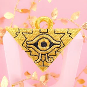 May include: A gold embroidered patch featuring a black eye within a triangle. The patch has a loop at the top for attaching.