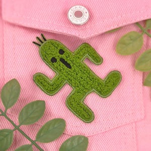 May include: A green cactus appliqué with black detailing is sewn onto a pink denim jacket pocket. The jacket has a silver button with "JEANS" on it. Green leaves are in the background.