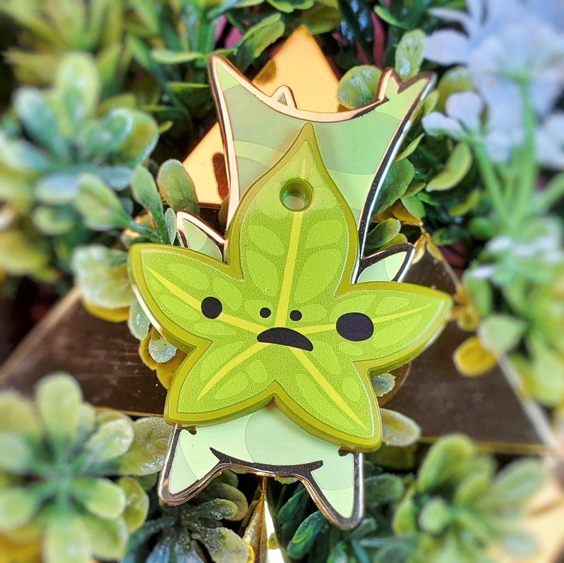 May include: A green leaf-shaped pin with a cartoon face and black eyes. The pin is made of metal and has a gold finish.