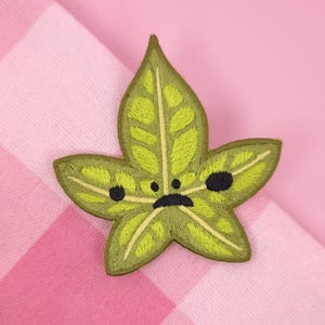 May include: Embroidered green leaf patch with a sad face, featuring black eyes and a curved mouth. The leaf has detailed stitching and a light green interior. The patch is set against a pink and white checkered background.