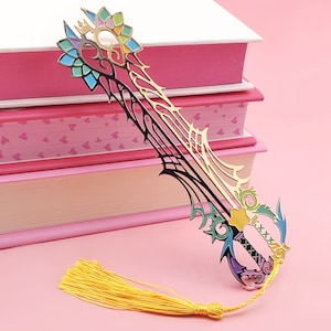 Puede incluir: Un marcapáginas de metal de colores con forma de llave espada y un borla. El marcapáginas está encima de una pila de libros con cubiertas rosas.