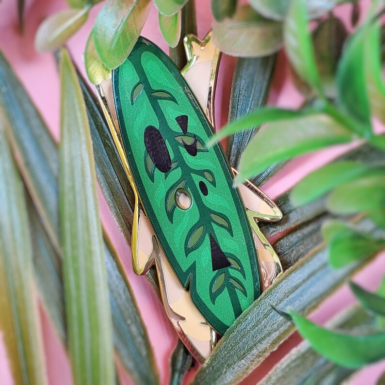 May include: A gold and green enamel pin featuring a stylized leaf design. The pin is surrounded by green foliage.