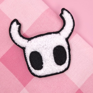 Peut inclure: Écusson brodé blanc et noir représentant une tête de personnage stylisée avec deux grands yeux ovales noirs et des cornes incurvées. L'écusson a une bordure noire et un aspect texturé et flou. Le fond est un motif à carreaux roses et blancs.