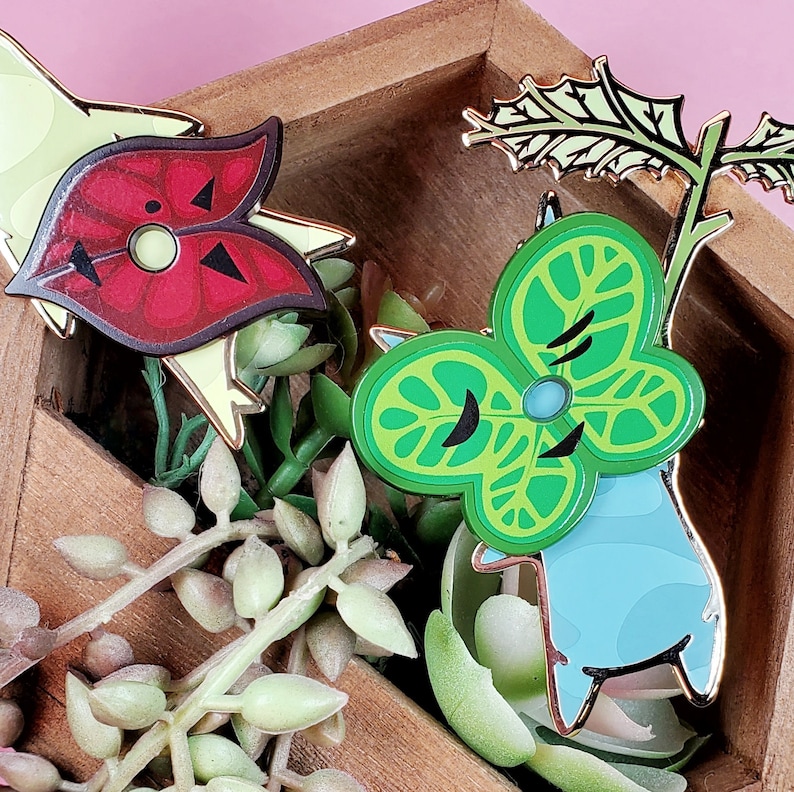 May include: Two enamel pins in a wooden box with green succulents. One pin is a red and yellow stylized heart with black markings. The other pin is a green and blue stylized leaf with a black eye.