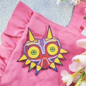 May include: A pink canvas tote bag featuring an embroidered Majora's Mask from the Legend of Zelda video game series.