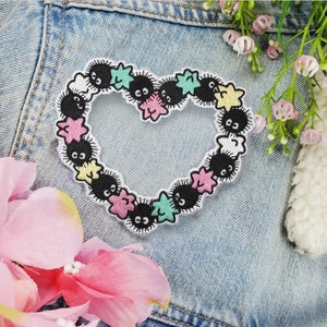 May include: An embroidered heart-shaped patch featuring a circle of black and white cartoon characters with stars in between. The characters have large eyes and are surrounded by a white outline.