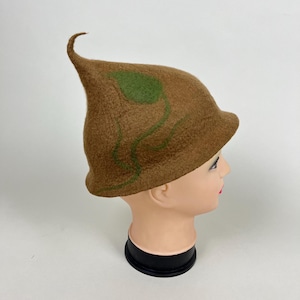 Op de afbeelding: Een bruine vilten hoed met een puntige top en een groen bladeren ontwerp. De hoed wordt getoond op een mannequin hoofd. Het bladeren ontwerp heeft groene stelen die langs de zijkant van de hoed naar beneden buigen.